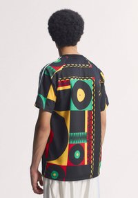 Άτομο με κοντά σγουρά μαλλιά που φοράει μια πολύχρωμη κοντομάνικη μπλούζα με αφηρημένο μοτίβο σε μαύρο, κόκκινο, κίτρινο και πράσινο, στραμμένο πλάτη σε λευκό φόντο.