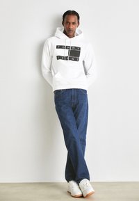 Felpa bianca con logo nero "TOMMY JEANS", abbinata a jeans in denim blu. Il modello indossa sneakers bianche con suola in gomma, mentre si trova davanti a un muro grigio.