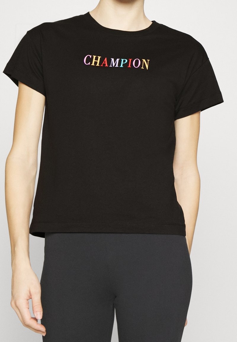 Personne portant un t-shirt court noir avec le mot "CHAMPION" en lettres multicolores sur la poitrine.