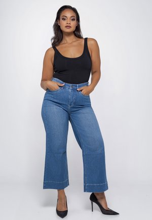 Vrouw staat met handen in zakken, draagt een zwarte mouwloze top, hoge taille wijde blauwe jeans en zwarte hoge hakken tegen een witte achtergrond.