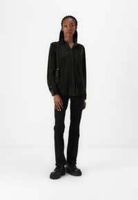 Camisa de botões de riscas verticais verde-escuro, feita de um tecido texturizado, combinada com calças pretas de perna reta e sapatos pretos robustos.
