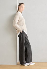 Mujer con el cabello recogido, vestida con un suéter beige, una camisa blanca, pantalones anchos negros y zapatillas blancas, de pie de perfil frente a una pared gris claro.