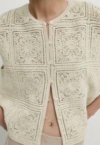 Beige virkad ärmlös topp med blommiga fyrkantiga mönster, små knappar framtill, bära över ljusa byxor, som visar midjan och händerna i fickorna.