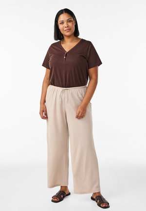 Kobieta stojąca, ubrana w brązową bluzkę z krótkim rękawem, beżowe spodnie o szerokich nogawkach z sznurkiem oraz czarne klapki, na białym tle.