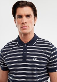 Tricou polo bleu marin cu dungi albe orizontale, placket cu două butoane și un mic logo brodat pe piept. Textură din material de bumbac.