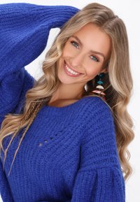 Maglione azzurro lavorato a maglia con un motivo testurizzato e un ampio scollo, dotato di maniche lunghe. Sono visibili anche grandi orecchini colorati.