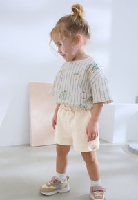 Chemise beige clair à manches courtes avec broderie de fruits colorés, associée à un short crème avec des éclats pastel. L'enfant porte des baskets multicolores.
