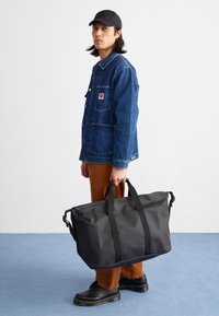 Sac de sport noir avec deux poignées noires, fabriqué en matériau résistant. Porté par une personne en veste en denim et pantalon marron, se tenant sur un sol bleu.