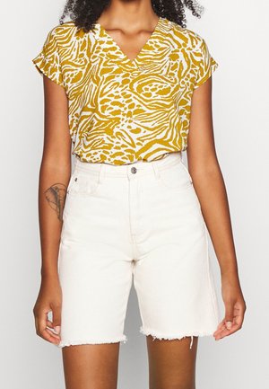 Kobieta o kręconych włosach, ubrana w musztardowo-biały wzorzysty T-shirt z dekoltem w serek, wpuszczony w białe, wysokie szorty jeansowe z postrzępionymi krawędziami.