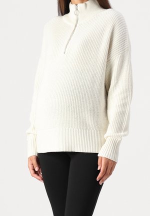 Femme portant un pull en tricot côtelé blanc avec un col à demi-fermeture éclair et des manches longues, associé à un pantalon noir.