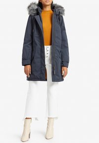 Navy parka with a removable faux fur hood, zip front, and side pockets, paired with a mustard top and white frayed hem jeans.