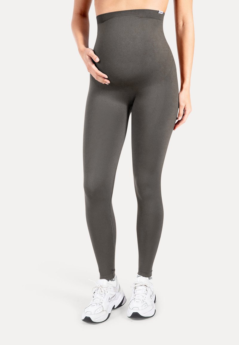 Personne enceinte portant des leggings de maternité gris foncé taille haute et des baskets blanches, main reposant sur le ventre, debout sur un fond uni.