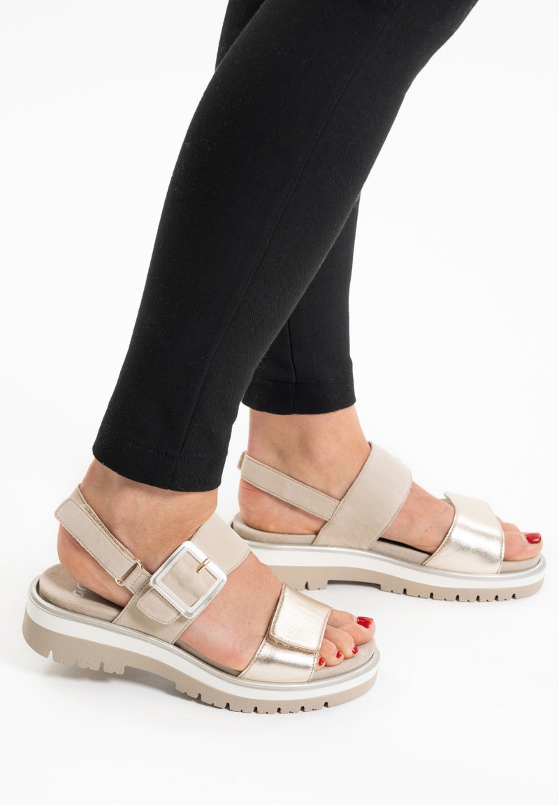 Pies con sandalias beige y doradas metálicas con hebilla y suelas gruesas, combinadas con leggings negros y esmalte de uñas de los pies rojo, sobre un fondo blanco.