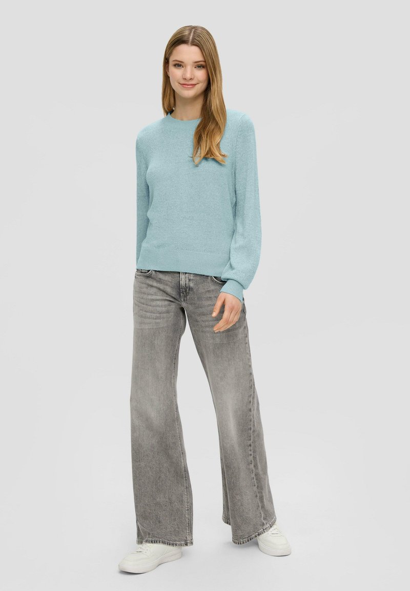 Helles blaues Strickpullover mit langen Ärmeln, kombiniert mit ausgewaschenen grauen Schlagjeans und weißen Sneakers, vor einem einfarbigen Hintergrund.