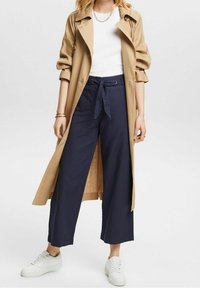 Brun trenchcoat med upprullade ärmar, över en vit t-shirt, kombinerat med marinblå vida byxor och vita sneakers. Silverkedjeaccessoar ingår.