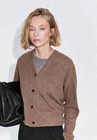 Bruine cardigan met een V-hals, lange mouwen en zwarte knopen. Gedragen over een grijze top, in combinatie met een zwarte tas. Soepele textuur.