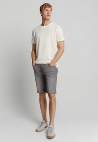 Heren lichtbeige textuur t-shirt, grijze knielange shorts en grijze sneakers met witte strepen. Model staat met de handen in de zakken.