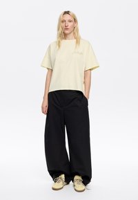 Maglietta cropped di un giallo chiaro con maniche corte e testo ricamato, abbinata a pantaloni larghi neri e scarpe di colore chiaro.