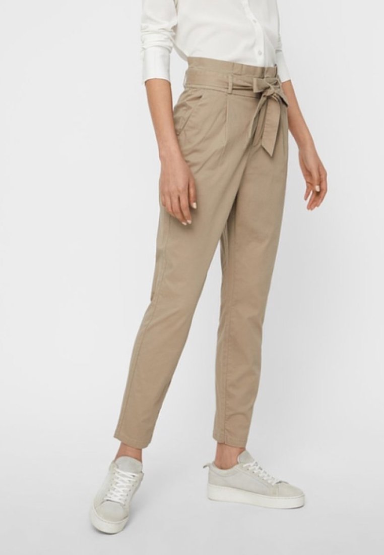 Pantalon kaki en mélange de coton, avec une taille haute, un détail de nouage à l'avant et des jambes fuselées. Porté avec une chemise blanche et des baskets.