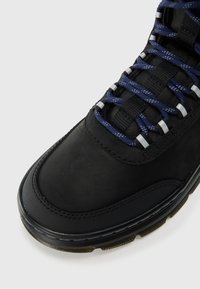 Chaussures noires avec un dessus en cuir lisse, une semelle en caoutchouc texturée, des lacets bleus et des détails argentés ; conçues pour la durabilité et le soutien.