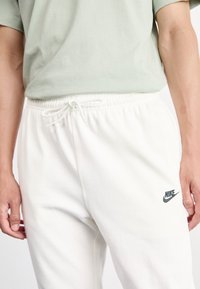 Pessoa vestindo calças de moletom brancas da Nike com cordão na cintura e uma camisa verde-claro, com as mãos relaxadas ao lado do corpo, mostrada da cintura até a metade da coxa.