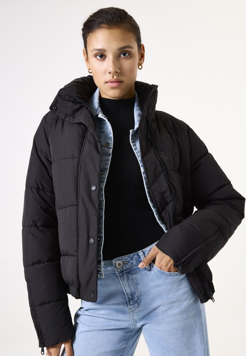 Schwarze Puffjacke mit Kapuze, ausgestattet mit einem Reißverschluss vorne und Druckknöpfen. Getragen über einem schwarzen Pullover und einem Denim-Hemd, kombiniert mit hellen Jeans.