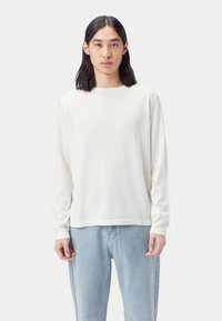 Weißes Langarm-T-Shirt aus weicher Baumwolle mit Rundhalsausschnitt, kombiniert mit hellblauen Jeans, vor neutralem Hintergrund gezeigt.