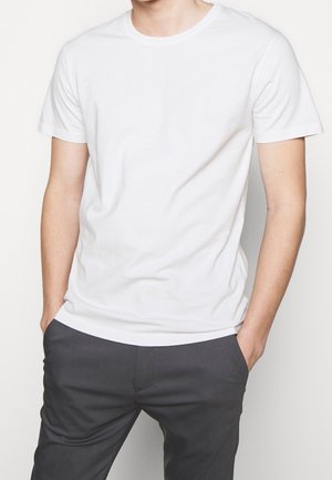 Weißes Baumwoll-T-Shirt mit Rundhalsausschnitt und kurzen Ärmeln, getragen mit dunkelgrauen Hosen. Glatte Textur und klassischer Schnitt.