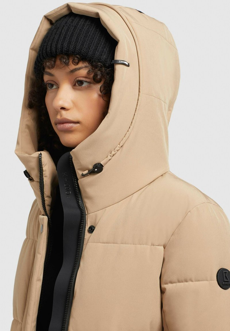 Femme portant une veste à capuche rembourrée beige par-dessus un bonnet en tricot noir, avec une fermeture éclair noire et des boutons-pression visibles sur la veste.