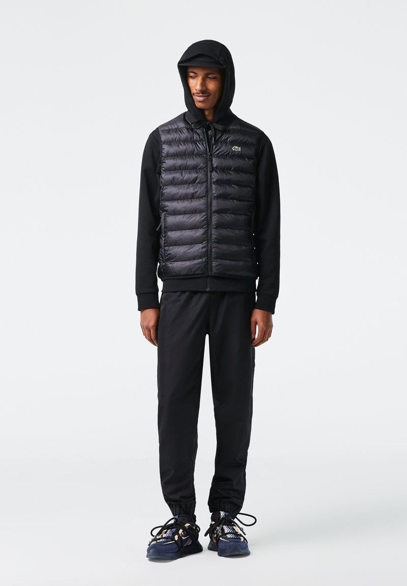 Un gilet matelassé noir sur un haut à manches longues noir, accompagné d'un pantalon noir. Des baskets colorées et texturées avec des lacets complètent la tenue.