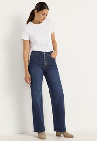 Farkut, joissa on korkea vyötärö, nappikiinnitys, suora lahje ja hieman haalistunut pinta. Yhdistettynä yksinkertaiseen valkoiseen t-paitaan.