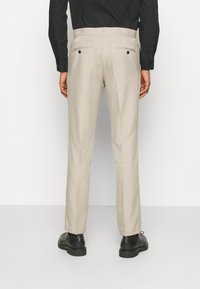 Pantaloni beige su misura con finitura in tessuto liscio, dotati di tasche posteriori e taglio dritto, abbinati a scarpe nere.