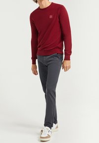 Bordowy sweter z dzianiny z okrągłym dekoltem, w połączeniu z szarymi spodniami slim fit i białymi trampkami z brązowymi akcentami.