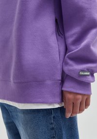 Sweat-shirt violet en mélange de coton, avec une fermeture éclair latérale, des poignets côtelés et un patch logo. Associé à un jean bleu clair.