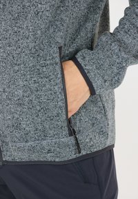 Grå sweater med en tekstureret stof, der har en lynlåslomme og en kontrastfarvet mørk kant langs kraven og manchetterne.