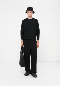 Sort sweatshirt og cargo-bukser kombineret med en bucket hat, mens der holdes en sort tote bag. Har en afslappet pasform og tekstureret stof.
