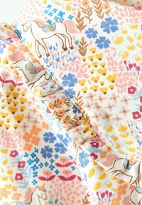 Biely textil s farebnými kvetinovými vzormi a malými koňmi s modrými sedlami, s volánovými okrajmi a jemnými ružovými, žltými a modrými kvetmi.