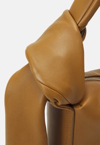Sac à main en cuir tan avec une finition douce et texturée ; il présente une poignée nouée, mettant en valeur des lignes épurées et des accents de design minimalistes.