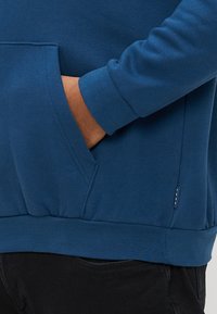 Hanorac albastru din bumbac, cu buzunar tip cangur, manșete și talie cu reiat, cu o etichetă mică pe lateral. Textură netedă.