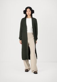 Lang, mørkegrønn strikket cardigan med ribbestruktur, åpen front og lange ermer, sammen med beige vidde bukser og svarte sko.