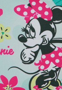 Minnie Mouse, ubrana w różową sukienkę w kropki i kokardkę, przyjmuje figlarną pozę z kolorowymi kwiatami w tle.