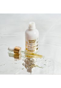 Bottiglia contagocce bianca con dettagli dorati, etichettata "Gradual Tan H2O Tan Drops", poggiata sull'acqua, con un tappo contagocce trasparente accanto.