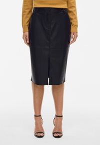 Γυναίκα φορώντας μία μαύρη δερμάτινη μίνι φούστα με άνοιγμα μπροστά, συνδυασμένη με ένα κίτρινο πουλόβερ και μαύρες γόβες με λουράκι στον αστράγαλο.