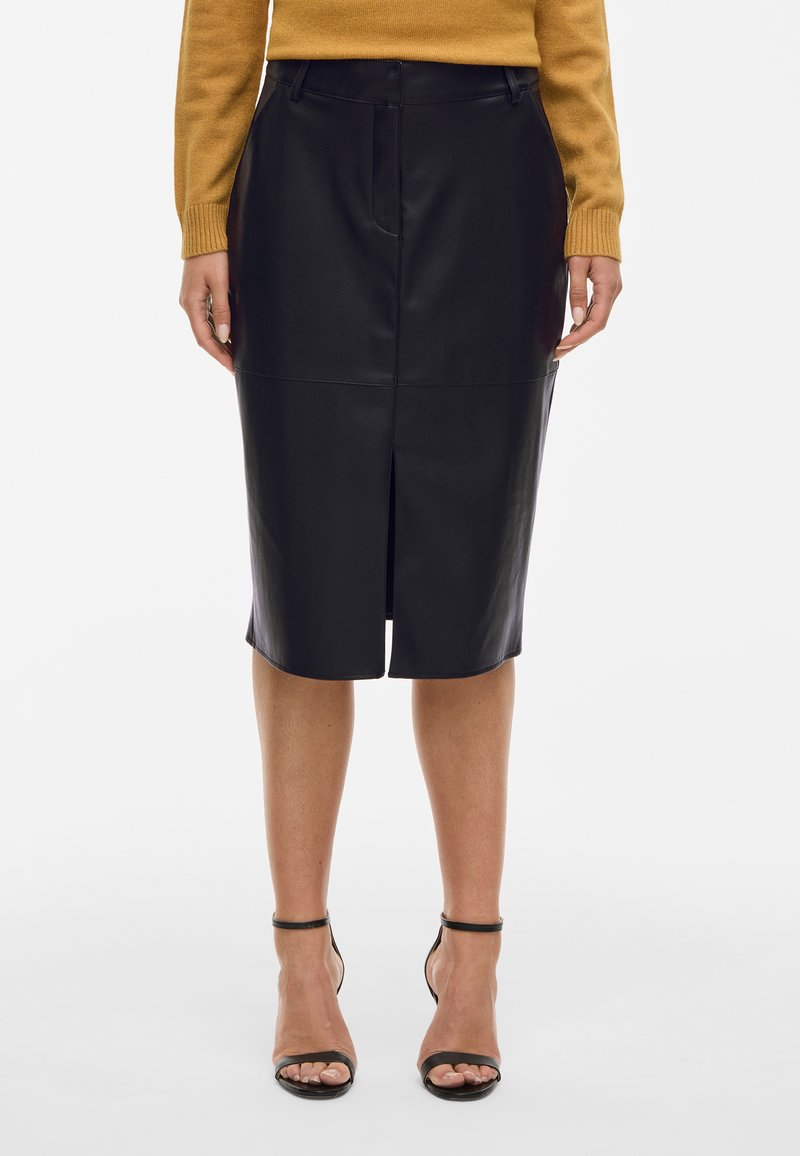 Γυναίκα φορώντας μία μαύρη δερμάτινη μίνι φούστα με άνοιγμα μπροστά, συνδυασμένη με ένα κίτρινο πουλόβερ και μαύρες γόβες με λουράκι στον αστράγαλο.