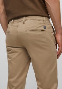 Béžové kalhoty vyrobené z hladkého materiálu, s dvěma zadními kapsami s horizontálními otvory a malou logo nášivkou na pravé straně.