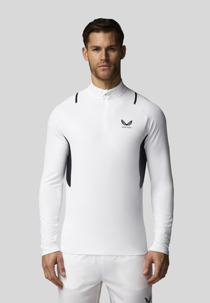 Homme portant un haut blanc ajusté à manches longues avec un col montant et un logo noir sur la poitrine, associé à un pantalon blanc, debout devant un fond gris.