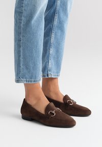 Mocassini in suede marrone con punta affusolata e dettagli in metallo argentato, abbinati a jeans cropped blu chiaro. Design con suola piatta.