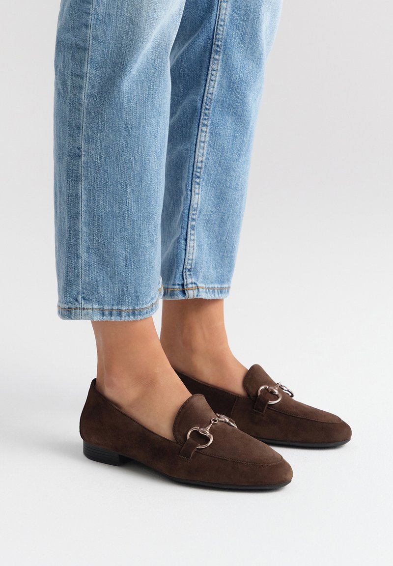 Mocassini in suede marrone con punta affusolata e dettagli in metallo argentato, abbinati a jeans cropped blu chiaro. Design con suola piatta.