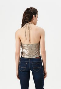 Zadnji pogled na zlato bleščeč halter crop top z vezanjem na vratu; v kombinaciji z temno modrimi kavbojkami z zadnjimi žepi in detajli šivanja.
