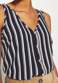 Top sans manches bleu marine avec des rayures blanches verticales, décolleté en V, boutons à l'avant, porté rentré dans un pantalon en velours côtelé tan avec taille boutonnée.